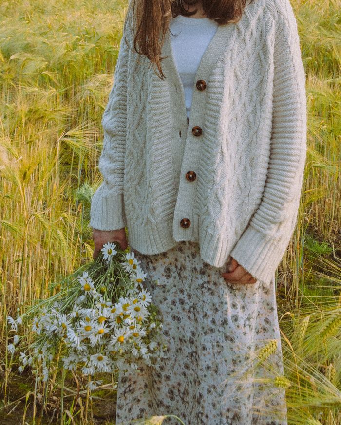 Wren cream cable-knit cardigan with tortoiseshell buttons, shown with a floral midi skirt and a bunch of white daisies in a barley field.