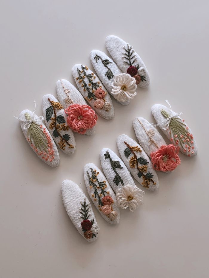 Posy handmade white linen snap hair clips embroidered with coral, cream and olive-green flowers, laid out in two diagonal rows.