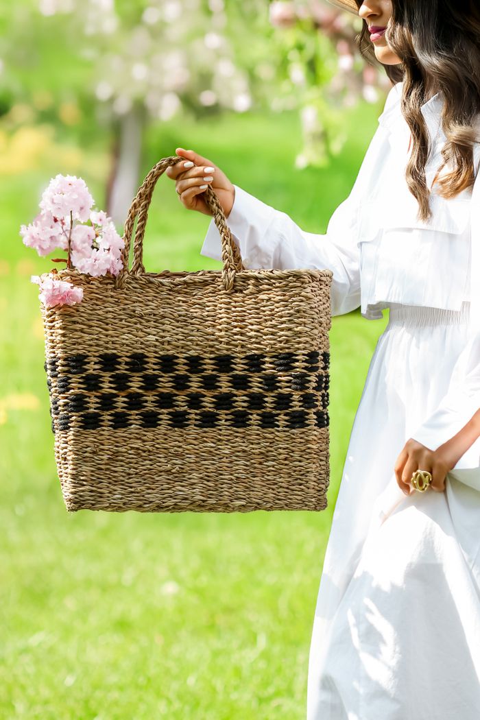 Meadow seagrass tote with a black woven band, filled with pink blossoms and carried across a green lawn.