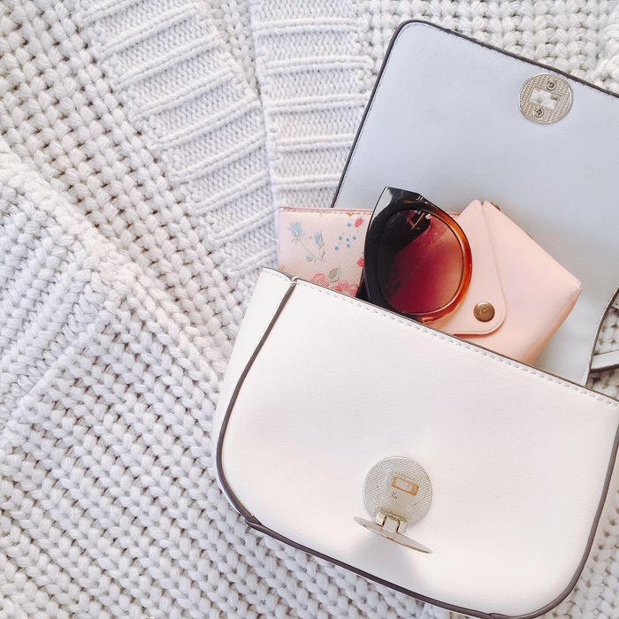 Daphne white leather crossbody bag with the flap open beside tortoiseshell sunglasses and a floral wallet, resting on chunky cream knitwear.