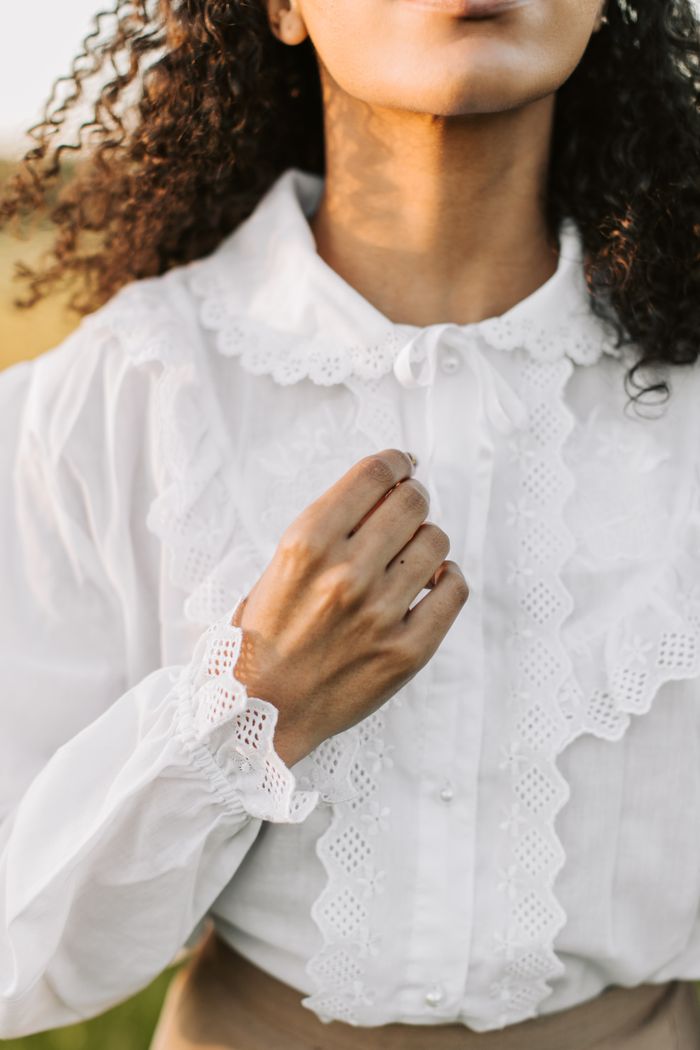 Odette white broderie blouse with a ruffled collar and cuffs, one hand resting at the chest.