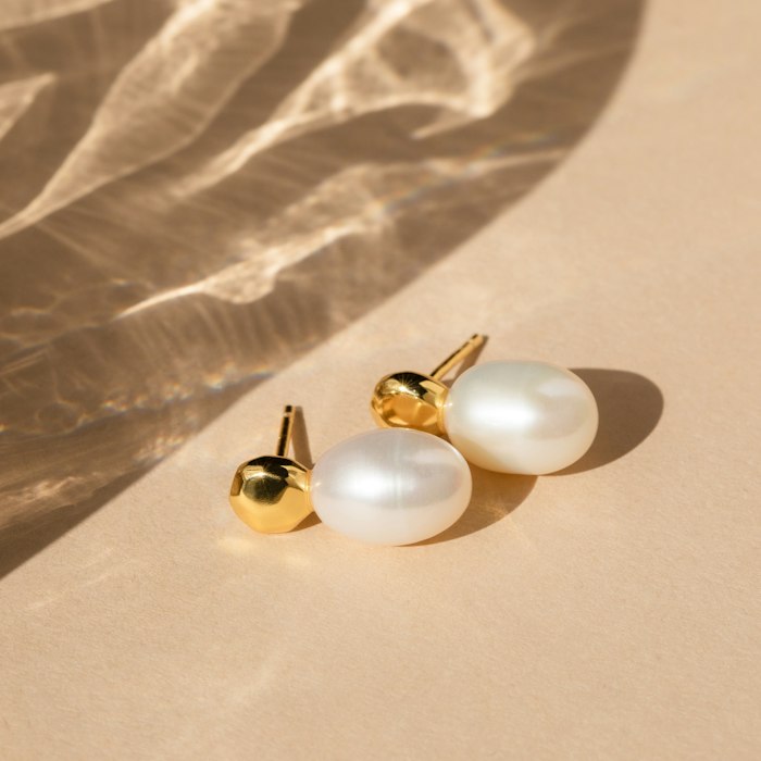 Perle gold-and-pearl stud earrings, each a faceted gold nugget with a large freshwater pearl drop, on a warm peach surface.