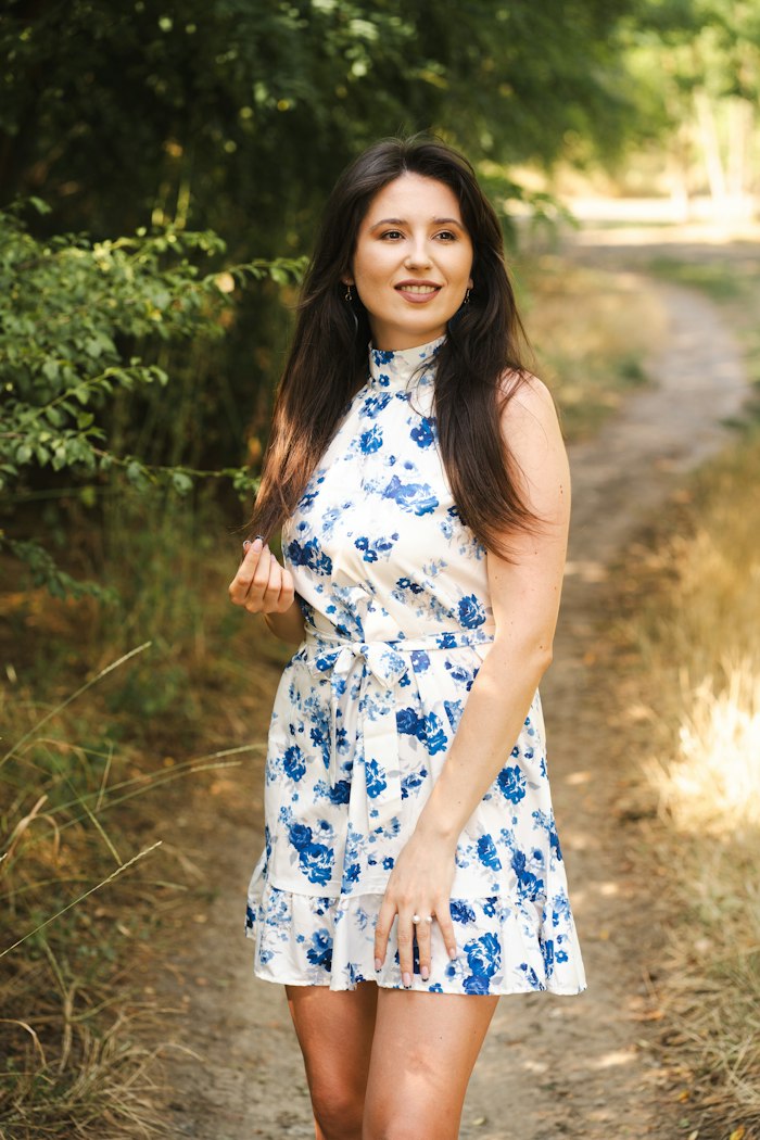 Marguerite halter dress in blue-and-white floral cotton voile with a tie waist and ruffled hem, worn on a sunlit woodland path.