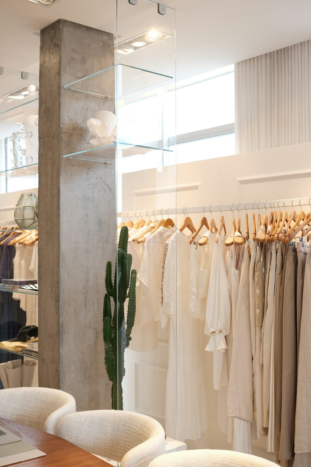 A bright minimalist boutique interior with a rail of cream and oat linen clothes on wooden hangers, a tall cactus, a concrete pillar and curved boucle chairs.