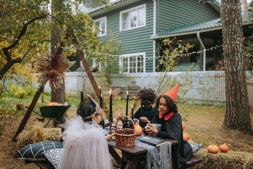 Three children in witch and masked costumes sit at a wooden table in a backyard decorated for Halloween, with pumpkins, hay bales, black candles and string lights between the trees.