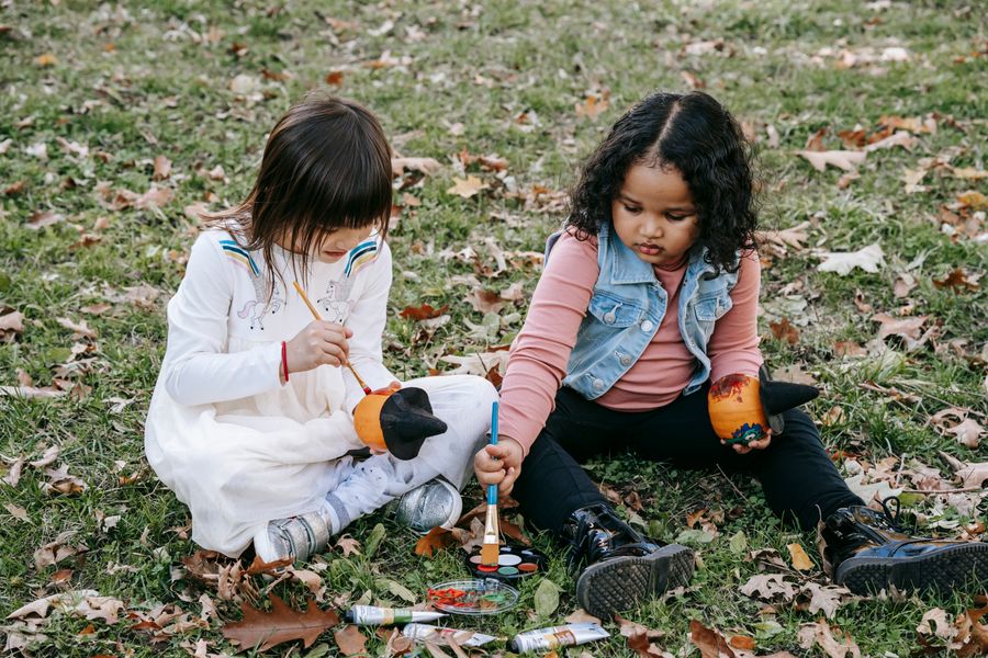 Two little girls sit on grass among fallen leaves, painting small pumpkins topped with black witch hats, with a paint palette and paint tubes beside them.