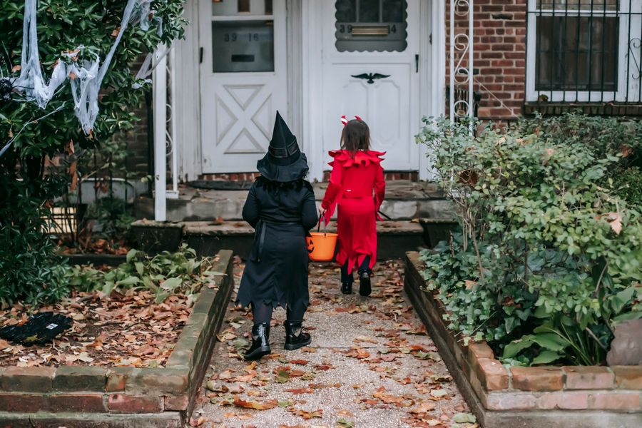 Two small children in a black witch costume and a red devil costume, seen from behind, walk up a leaf-strewn brick path with a jack-o-lantern bucket toward a white front door with cobwebs on the hedge.