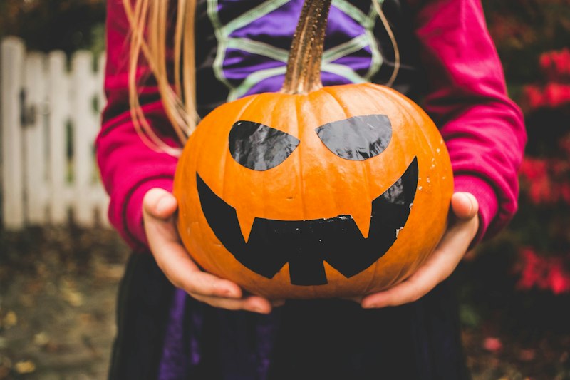 A child in a magenta top and purple costume holds a large pumpkin decorated with a grinning black vinyl face in both hands.