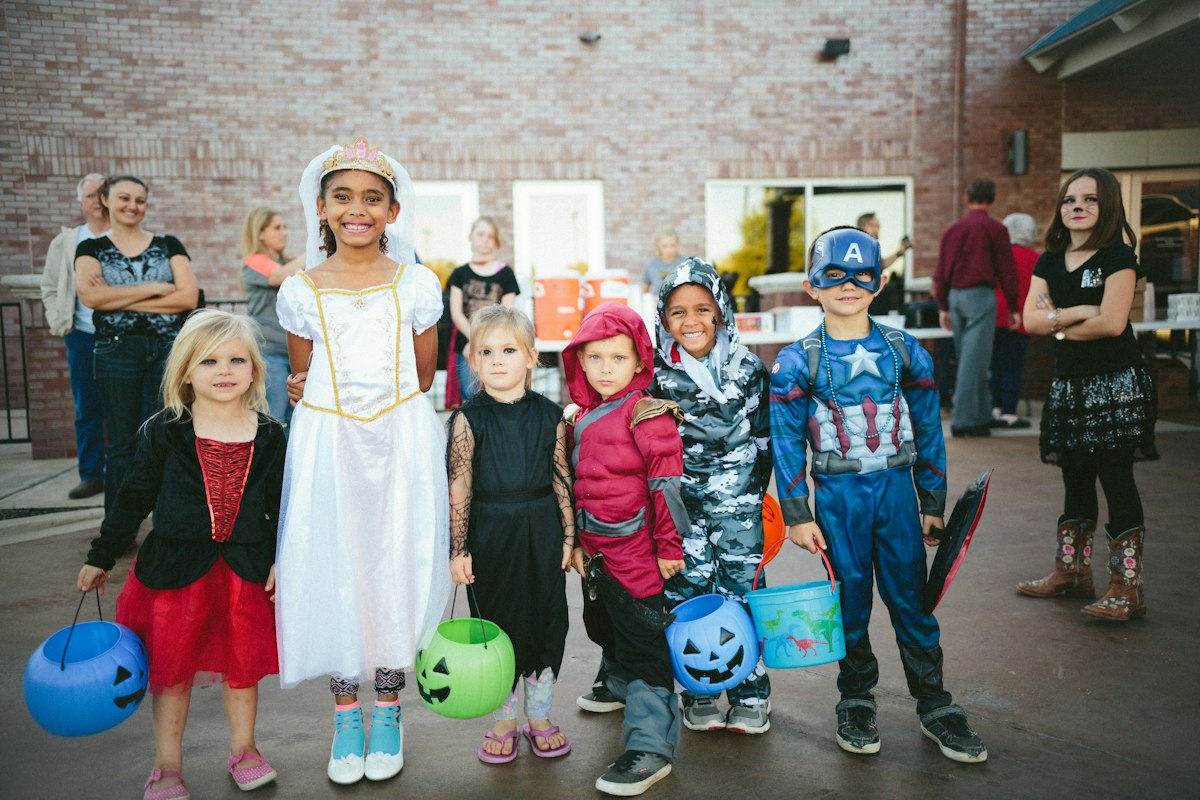 A row of small children in Halloween costumes, including a princess in white, a hooded ninja and a superhero, hold jack-o-lantern buckets in front of a brick building.