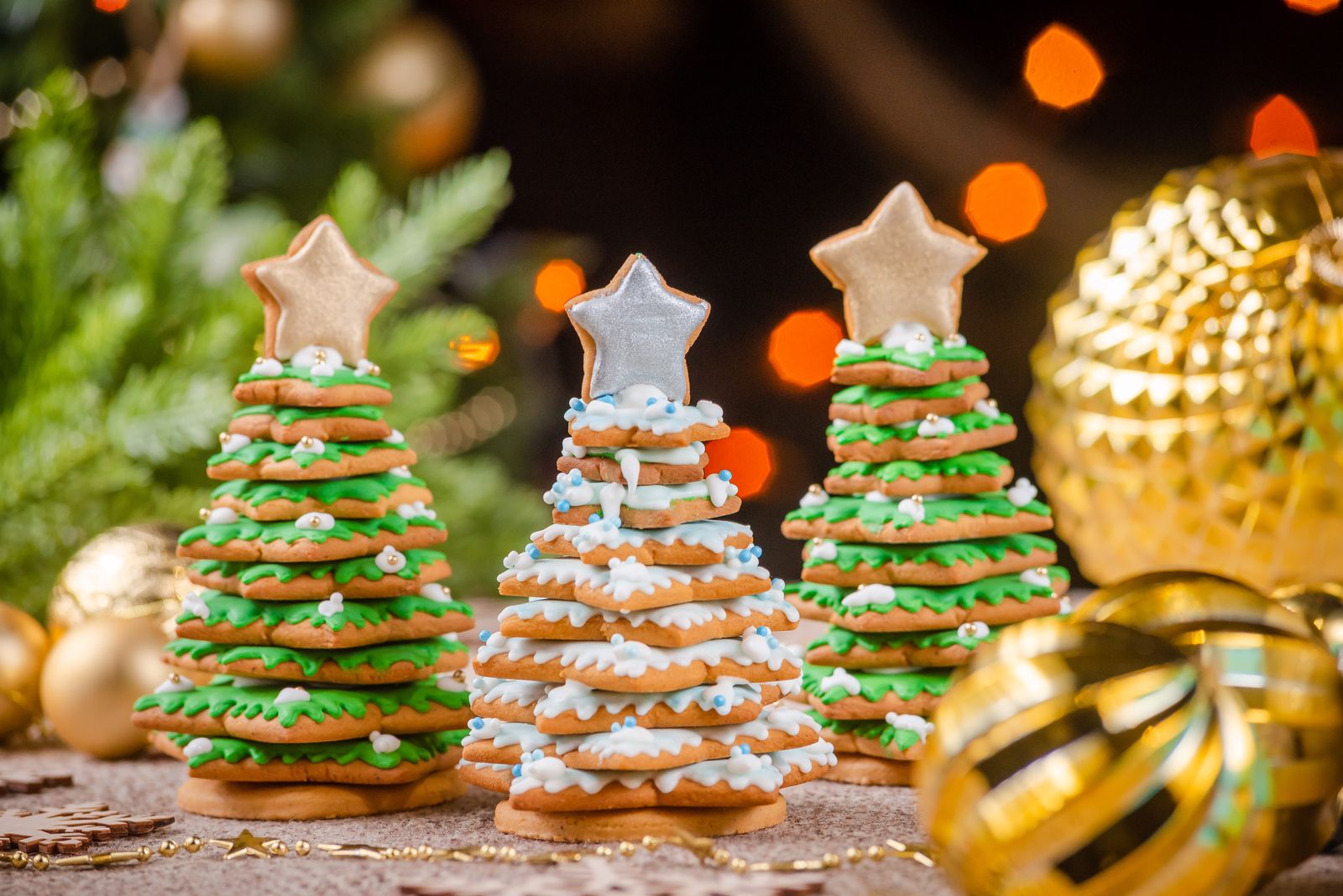 Three stacked gingerbread Christmas-tree cookies with green and white icing and star toppers, among gold baubles and warm blurred tree lights.