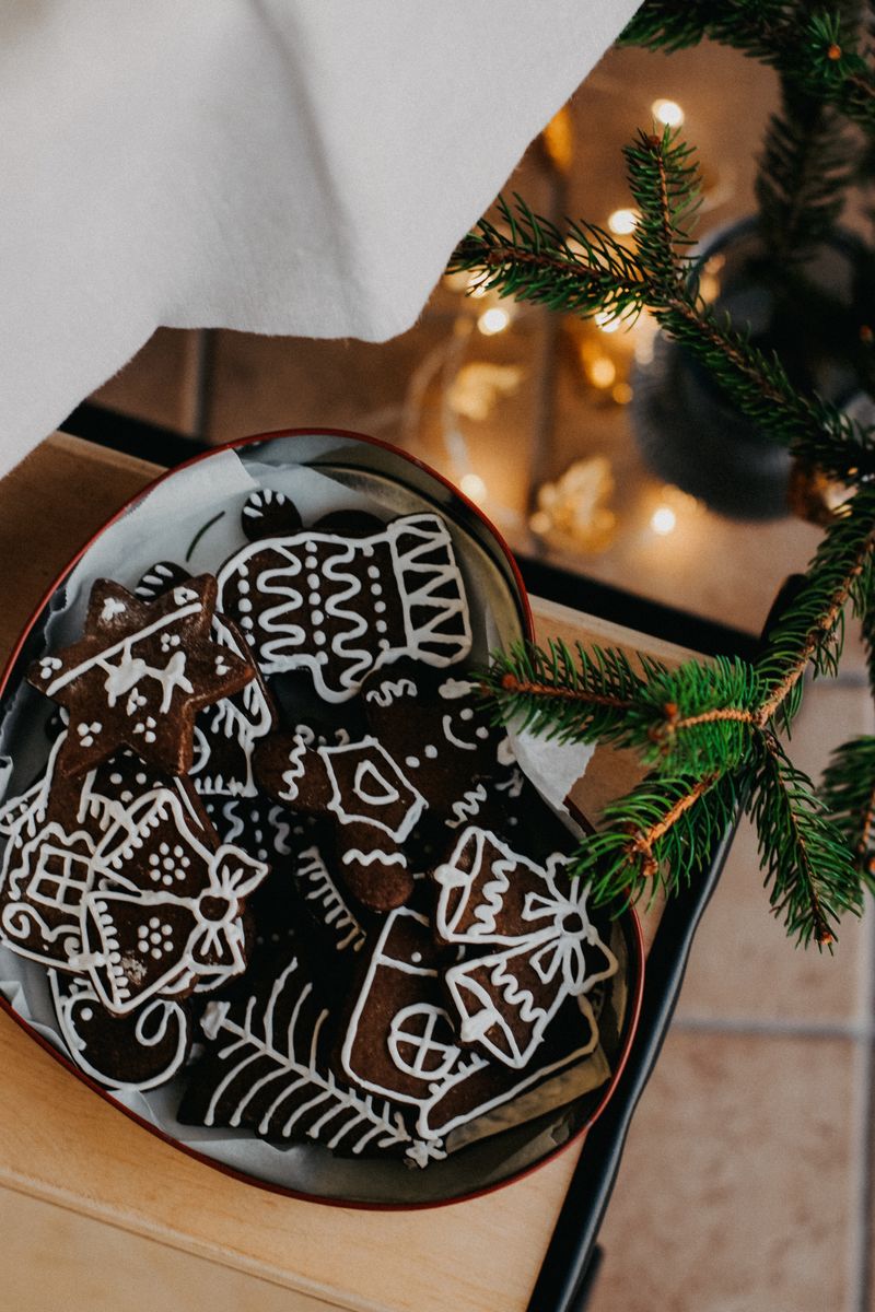 Overhead view of a red-rimmed oval tin lined with baking paper and packed with dark gingerbread cookies iced in white, beside a fir sprig with warm fairy lights glowing behind.