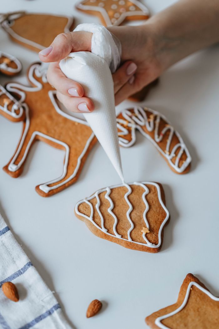 A hand piping white icing onto a gingerbread cookie, with other iced reindeer and star cookies on a pale table.