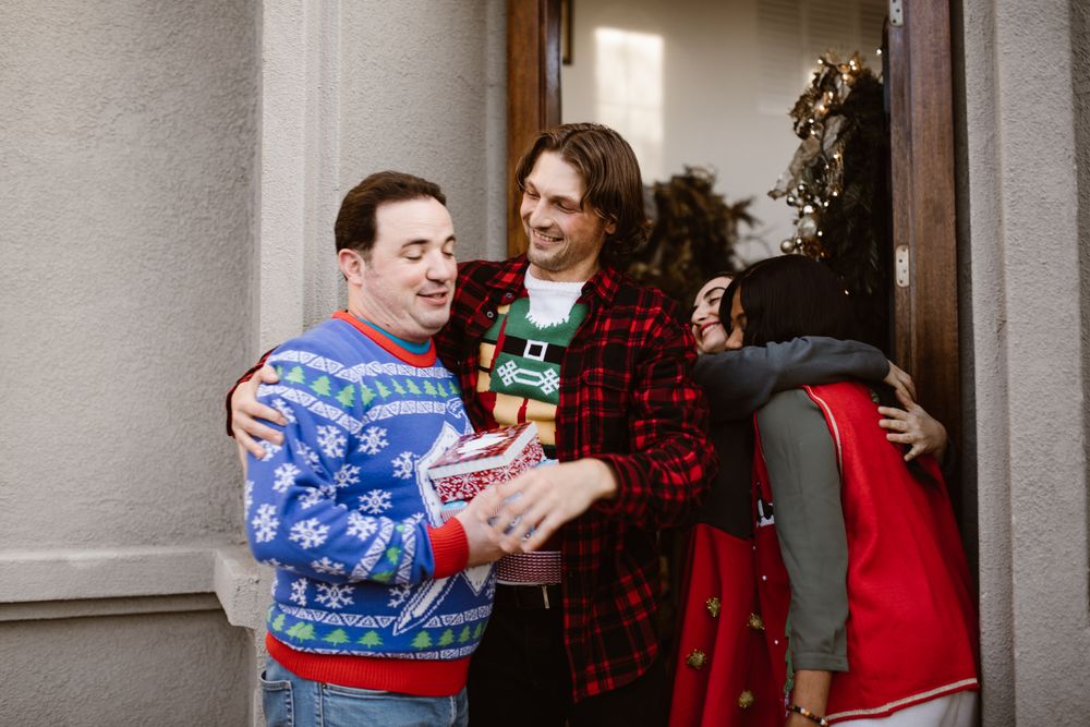 Friends in colourful ugly Christmas sweaters laughing and hugging in a doorway, one holding a small wrapped gift box, with a baubled garland on the door beside them.