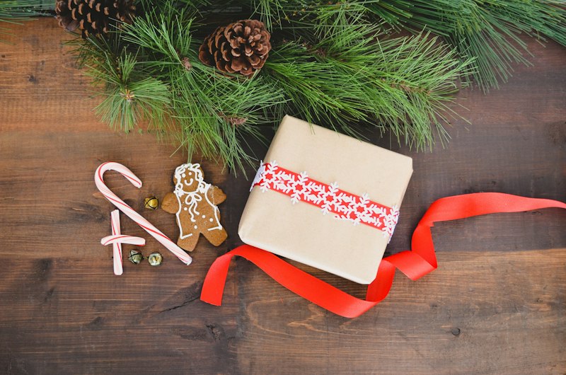 A gift wrapped in kraft paper with a red patterned band and ribbon, beside a candy cane, a gingerbread man and pine branches on a dark wooden table.