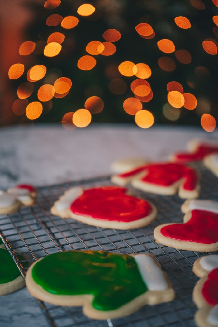 Red and green iced mitten and boot cookies on a wire cooling rack, with glowing orange tree lights out of focus above them.
