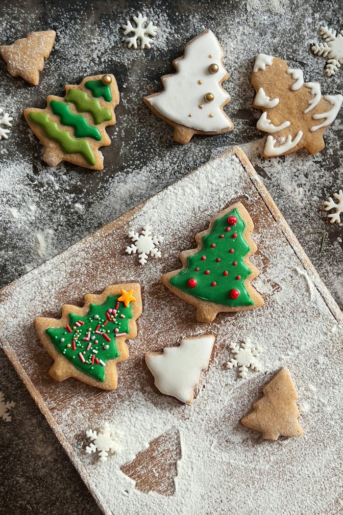Iced Christmas-tree cookies in green, white and gold, dusted with flour and scattered with sugar snowflakes on a light wooden board and a dark slate surface.