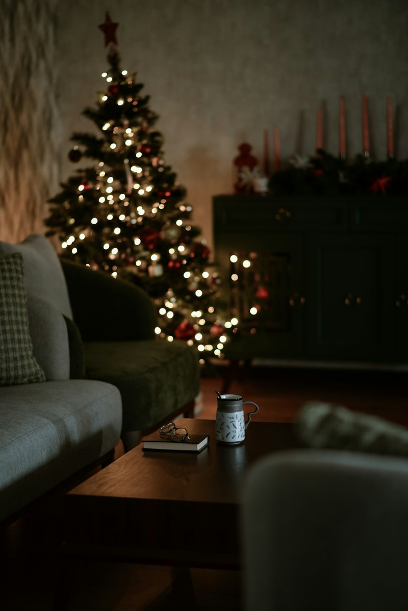 A dim, cosy living room with a softly lit Christmas tree glowing behind a sofa and a coffee table with a mug and a book.
