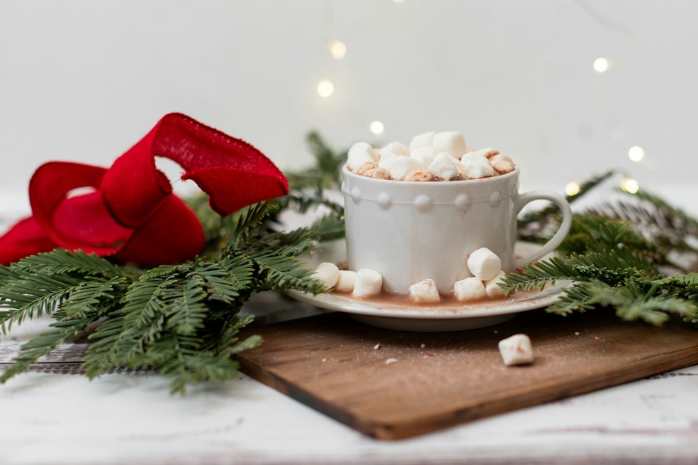 A white ceramic cup of hot chocolate piled with mini marshmallows on a wooden board, beside fir sprigs and a red ribbon, with soft fairy lights behind.
