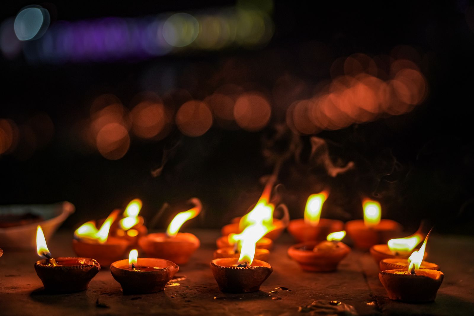 Small clay diyas burning with tall flames in loose rows along the bottom of a dark frame, with soft amber and violet bokeh lights and wisps of smoke above.