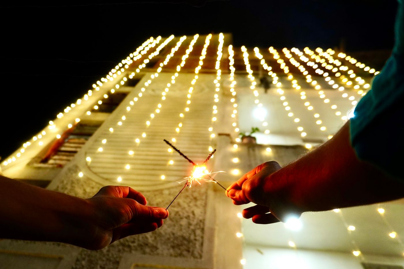 Two hands, one reaching in from each side, touch lit sparklers together in front of a building outlined with rows of warm fairy lights at night.