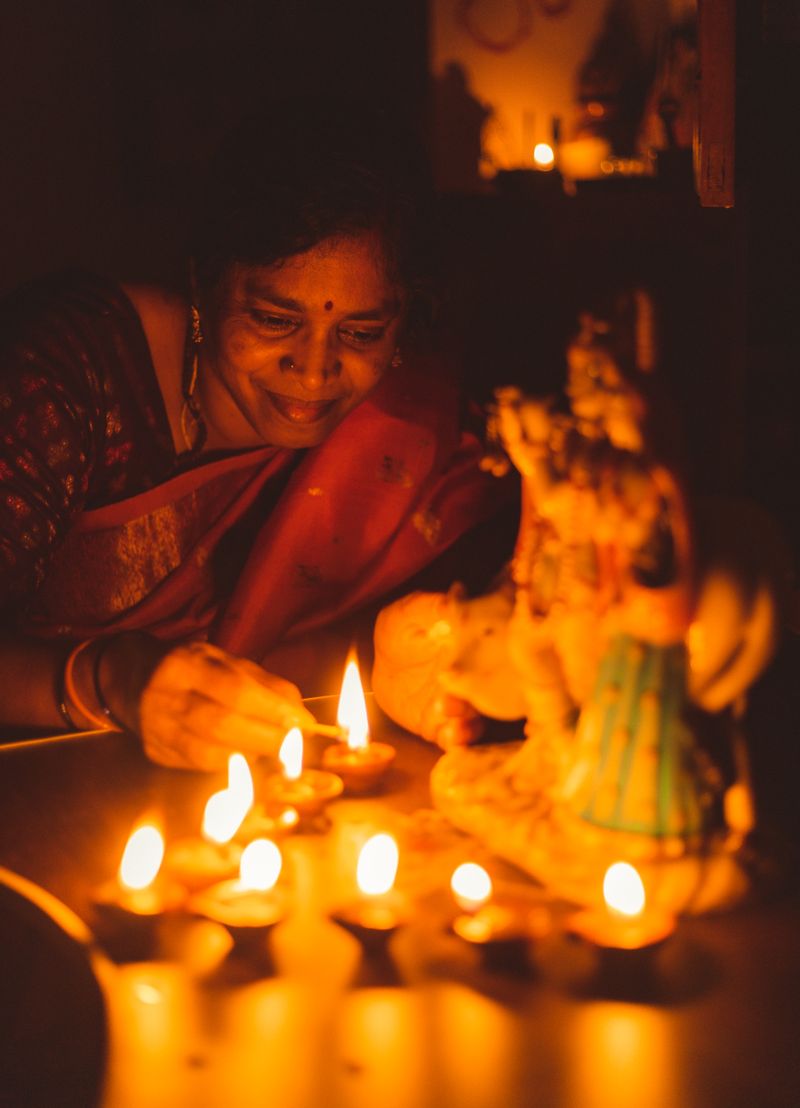 A smiling woman in a red sari lights small clay diyas in front of a blurred deity figure, her face lit by the warm flames in a dark room.