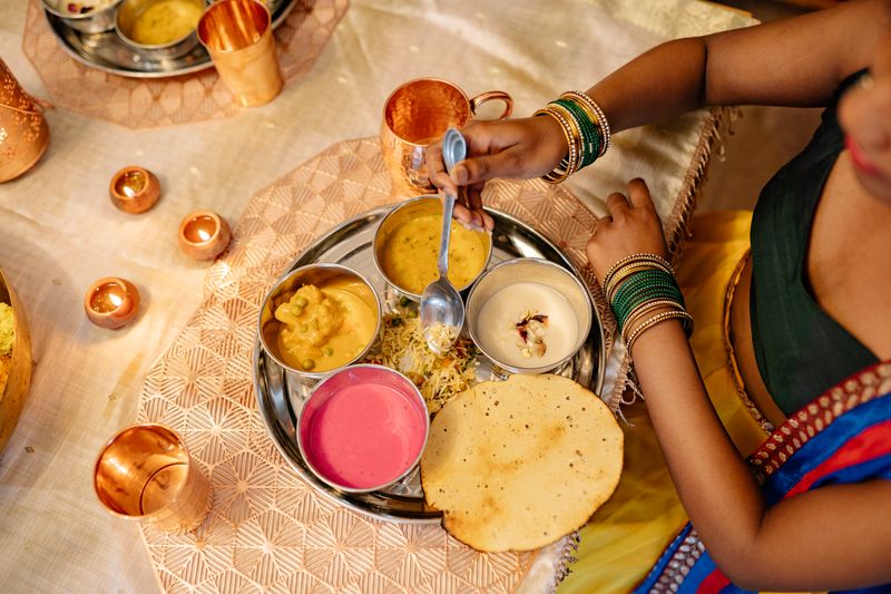 Overhead view of a woman in a sari spooning curry from a steel thali that also holds a pea curry, a white creamy sweet, a pink beetroot raita, rice and a papad, on a cream table with copper tumblers and small lit diyas.