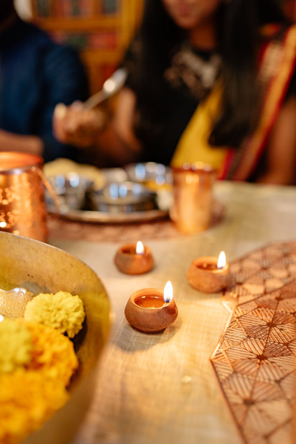 Small lit clay diyas and a bowl of yellow marigolds on a table where a Diwali meal is being served, with blurred guests in the background.