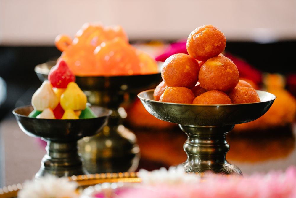 Antique brass pedestal bowls holding bright orange ladoo and colourful modak sweets, with pink and white flowers softly blurred in front.