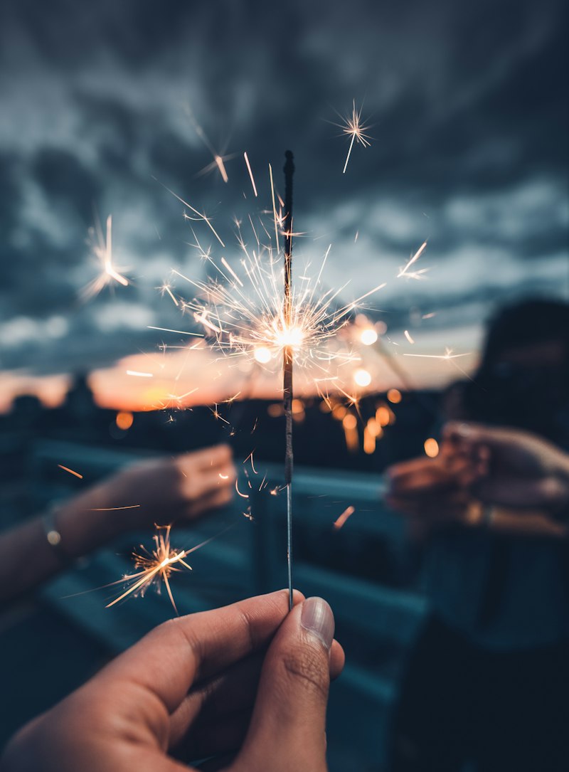 A hand holds a lit sparkler throwing fine sparks against a dusky blue and orange sky, with other hands blurred in the background.