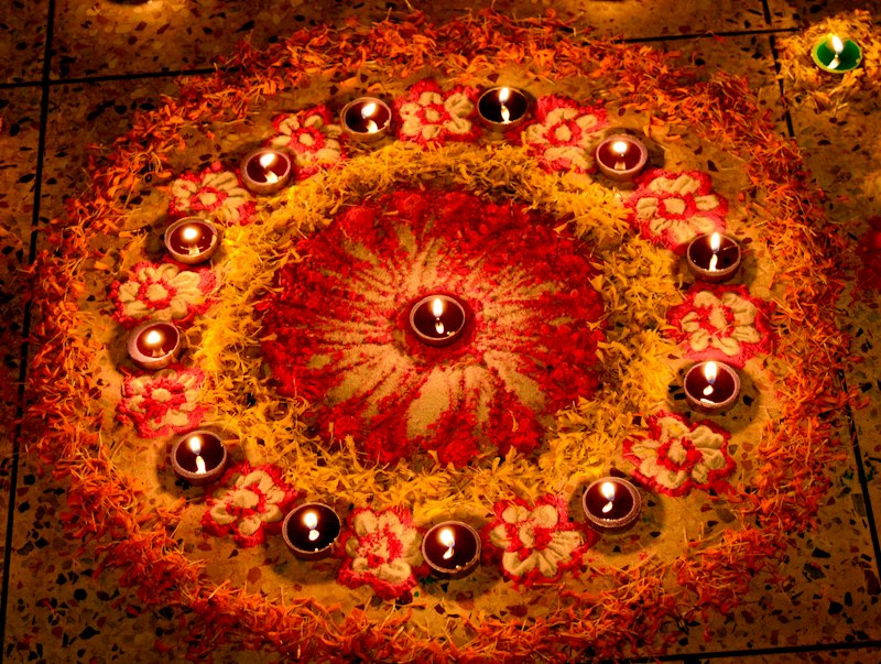 Top-down view of a circular floral rangoli on a tiled floor, built from red powder, orange and yellow marigold petals and pink flower motifs, ringed by small lit diyas.