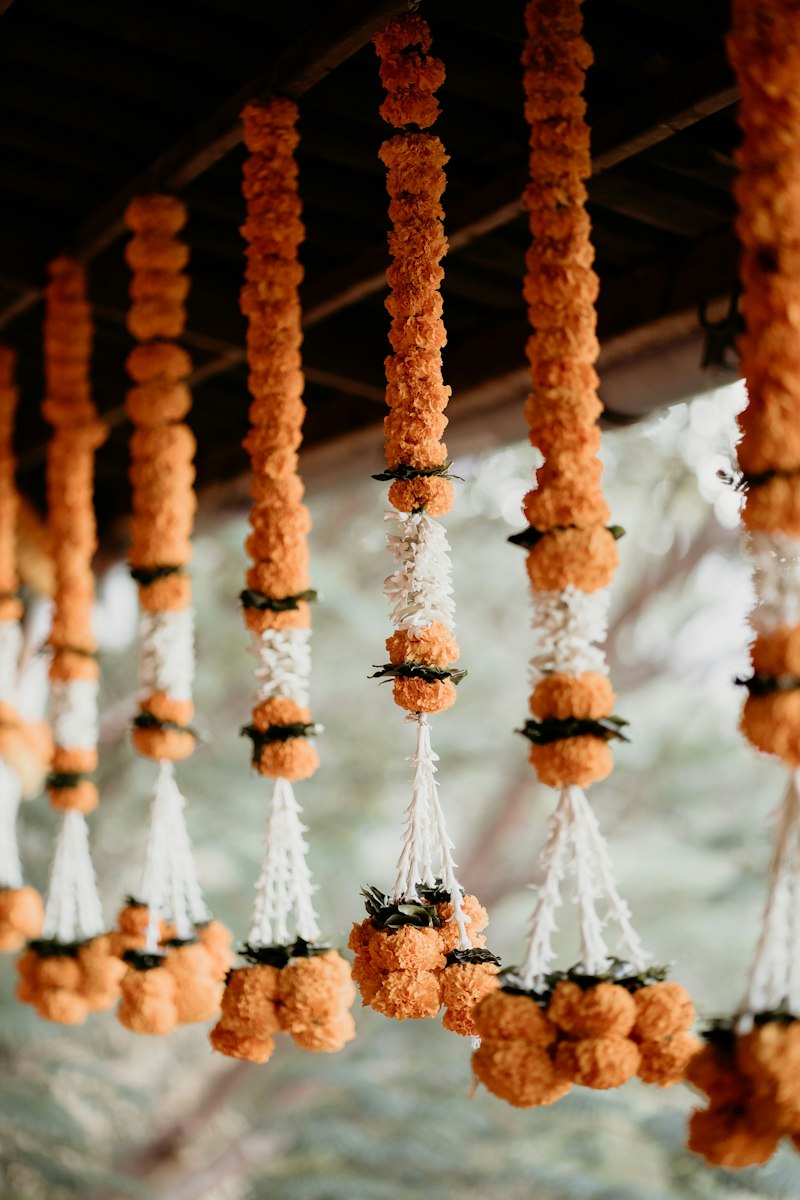 Hanging strands of orange marigolds with white jasmine, ending in white tassels and marigold clusters, under a dark wooden roof against a softly blurred green-grey background.
