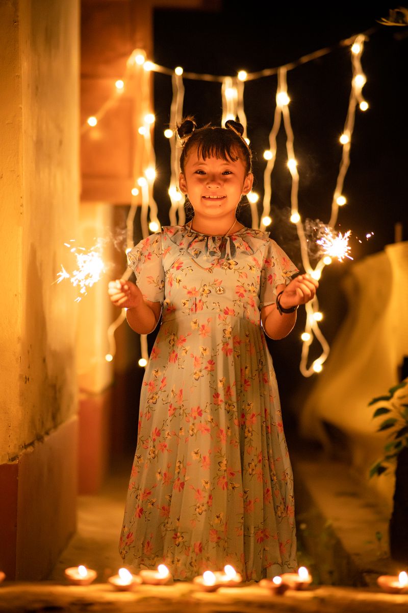 A little girl in a floral dress holds two sparklers at night in a lane strung with fairy lights, with small diyas glowing at her feet.