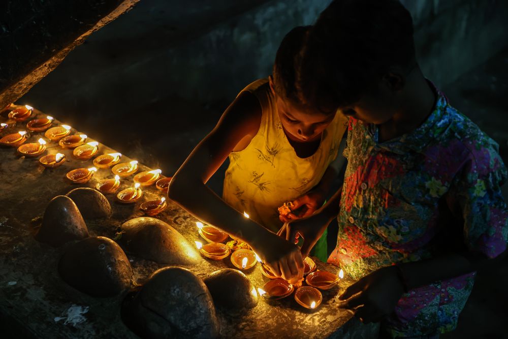 Two children bend over a long row of small lit diyas set along a wall at night.