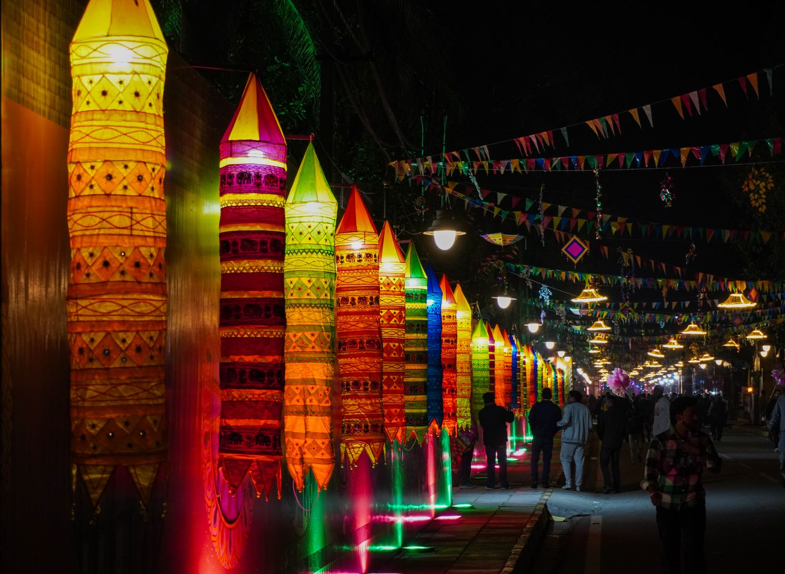 Tall glowing paper lanterns in orange, yellow, green and pink line a night street under strings of festival bunting while people stroll toward the lit end of the road.