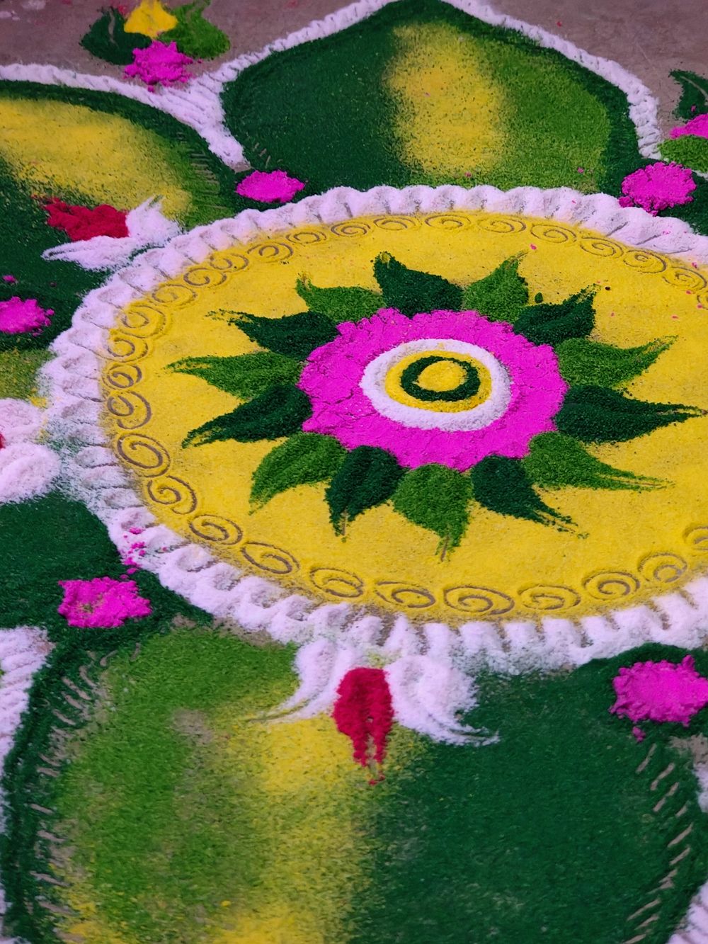Close view of a coloured-powder rangoli on the ground: a yellow medallion with a magenta flower and green leaves, ringed by white scrollwork and big green petals.