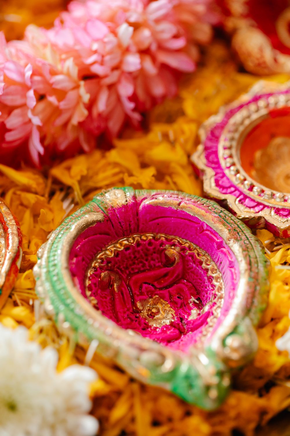 A painted clay diya in pink, green and gold sits among marigold petals and pink flowers.