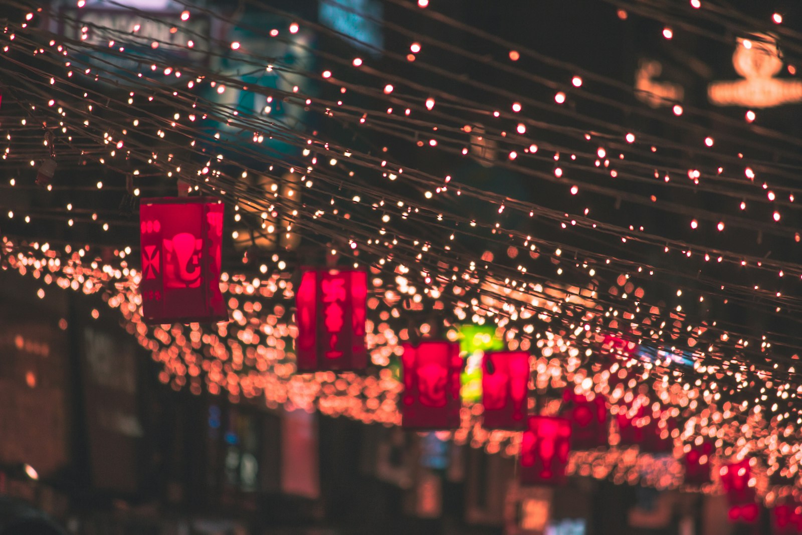 Strings of fairy lights stretch across a market lane at night with red paper lanterns hanging beneath them.
