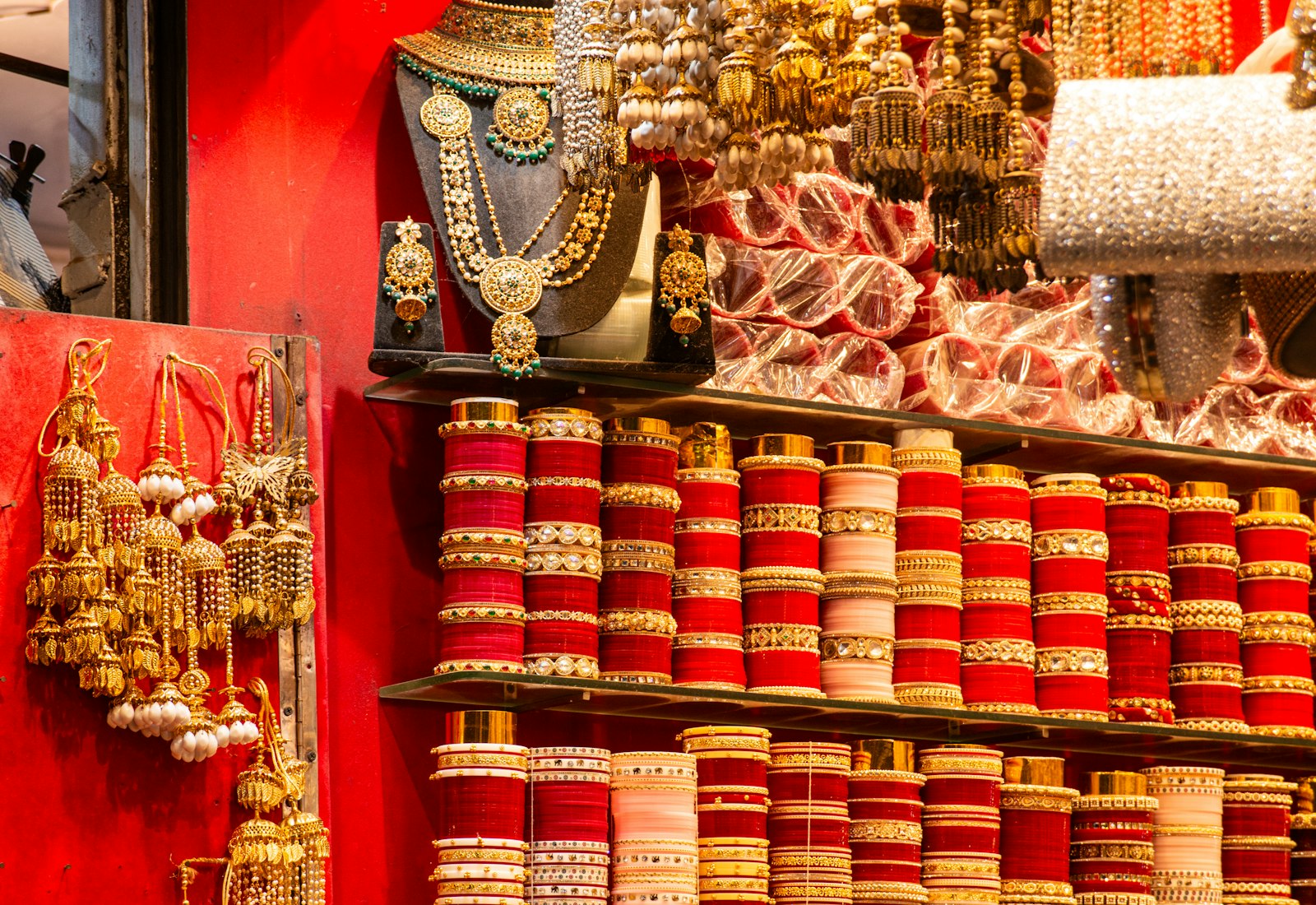 Market shelves stacked with red and ivory bangles beside gold tassels and necklaces against a red wall.