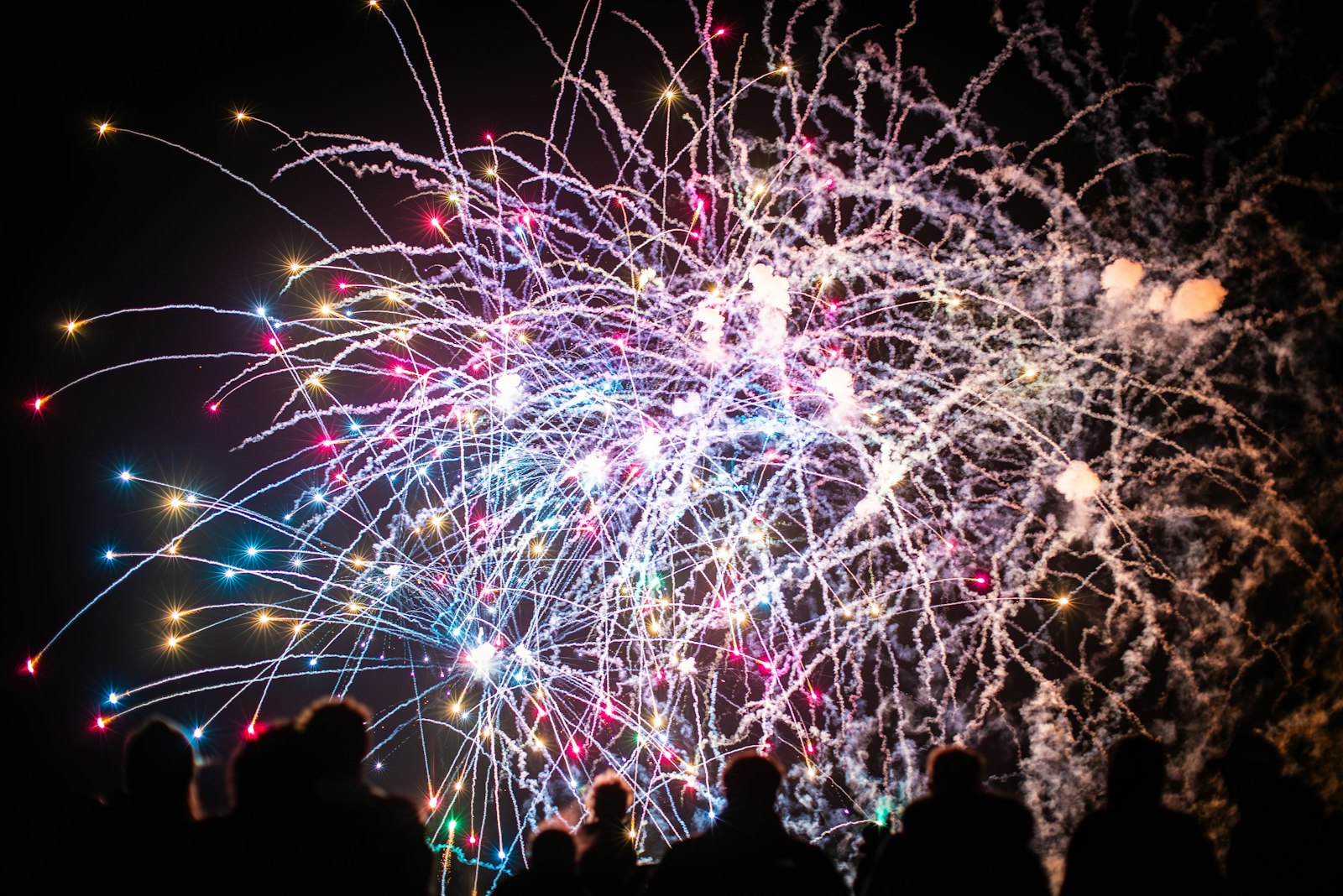 Multicoloured fireworks burst across a black night sky above a silhouetted crowd watching.