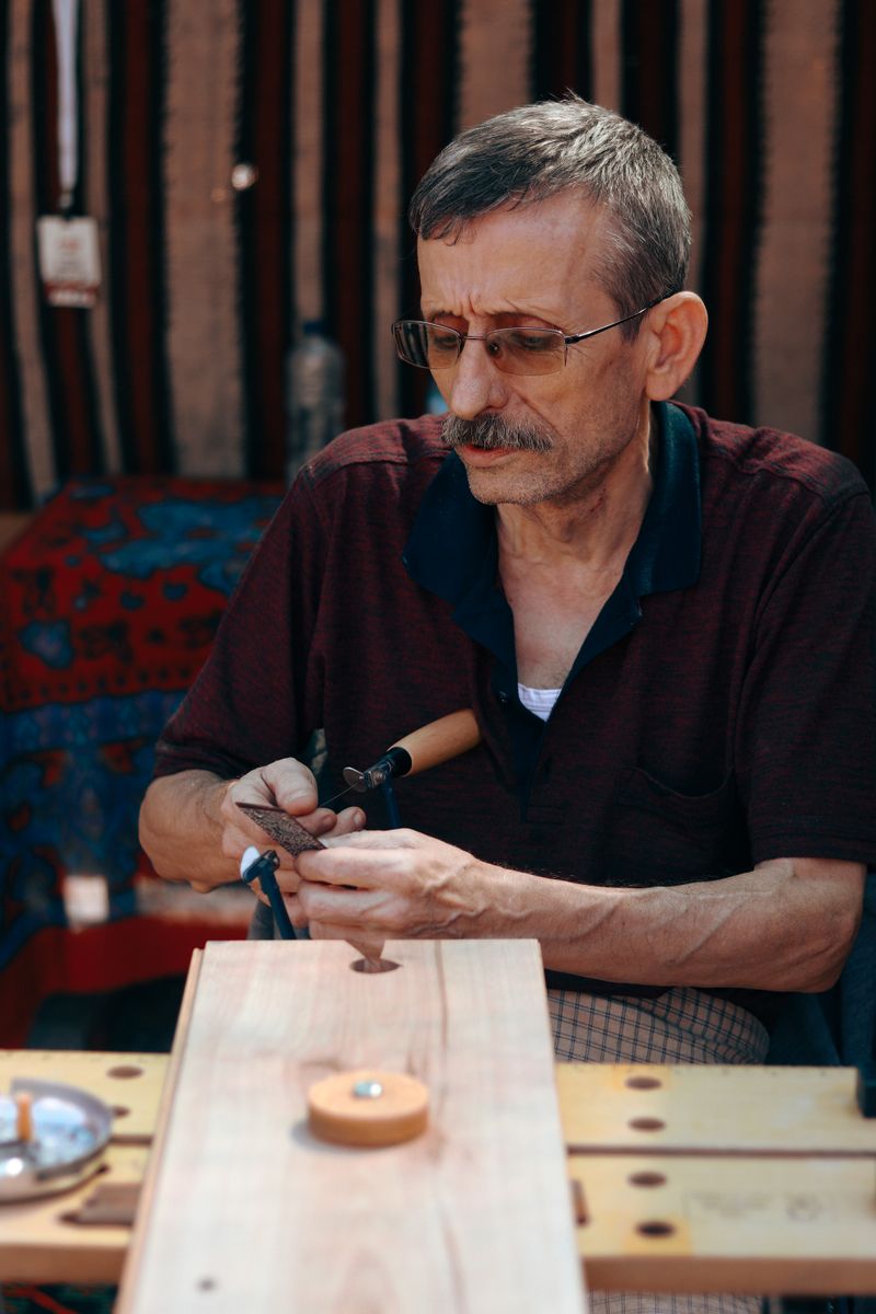 A gray-haired man in glasses and a maroon polo shirt concentrates on fine woodwork with a small fretsaw, a plank clamped on the bench in front of him.