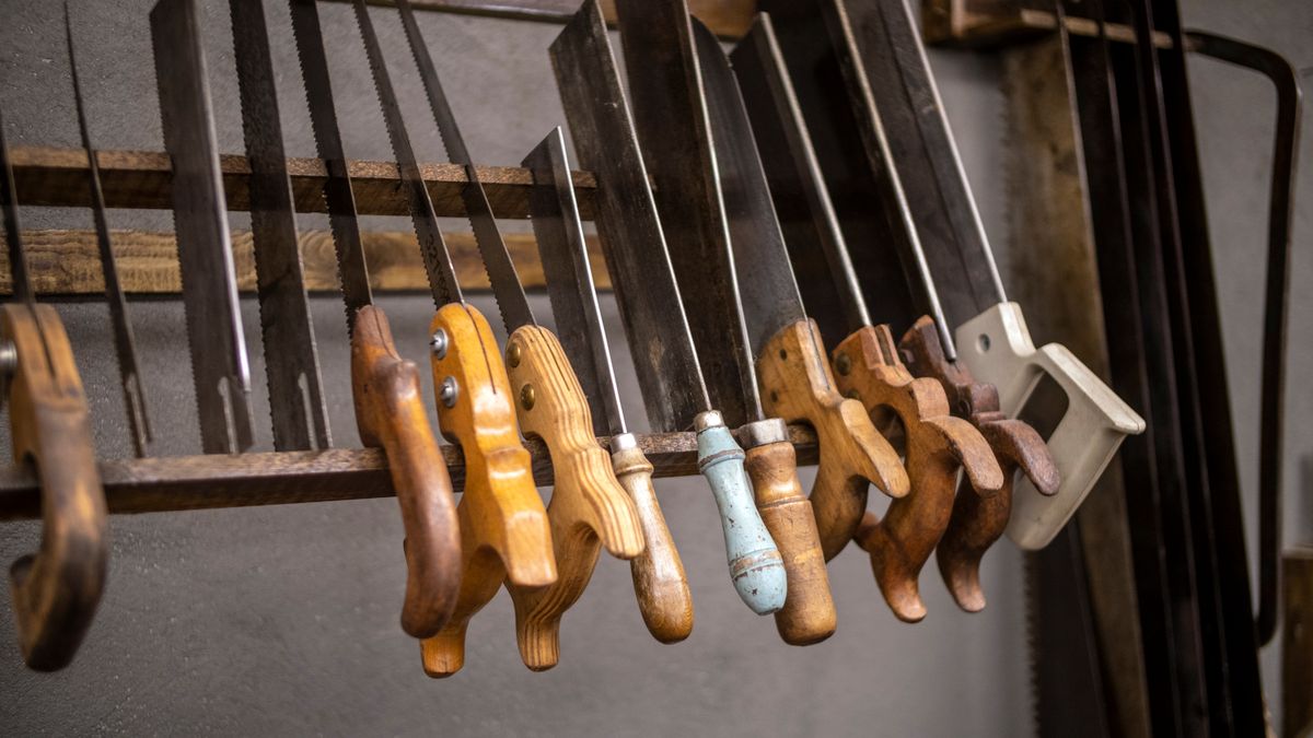 A row of vintage hand saws with worn wooden handles, blades upright, resting side by side on a wooden rack against a gray wall.