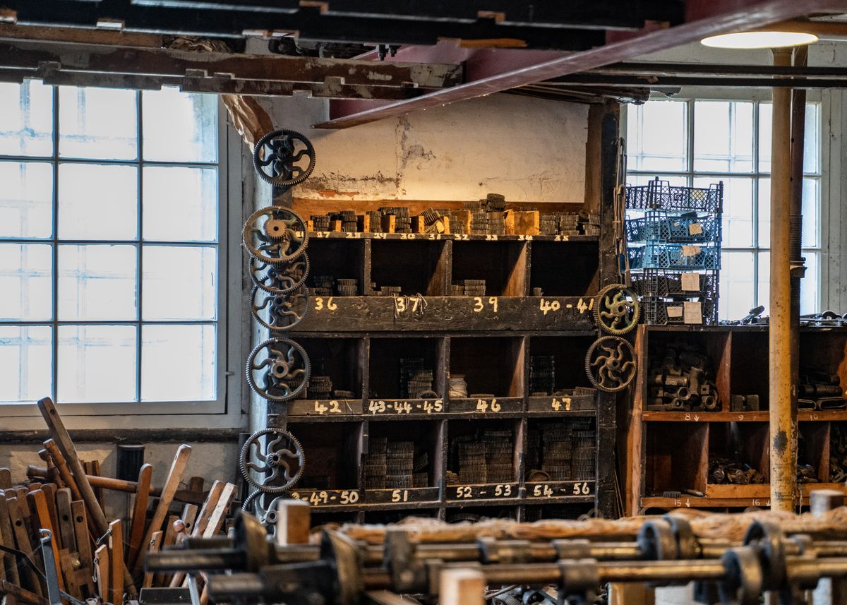 The back wall of an old hardware store: tall dark wooden pigeonhole shelves of hand-numbered bins, iron gear wheels on the side and daylight through large paned windows.