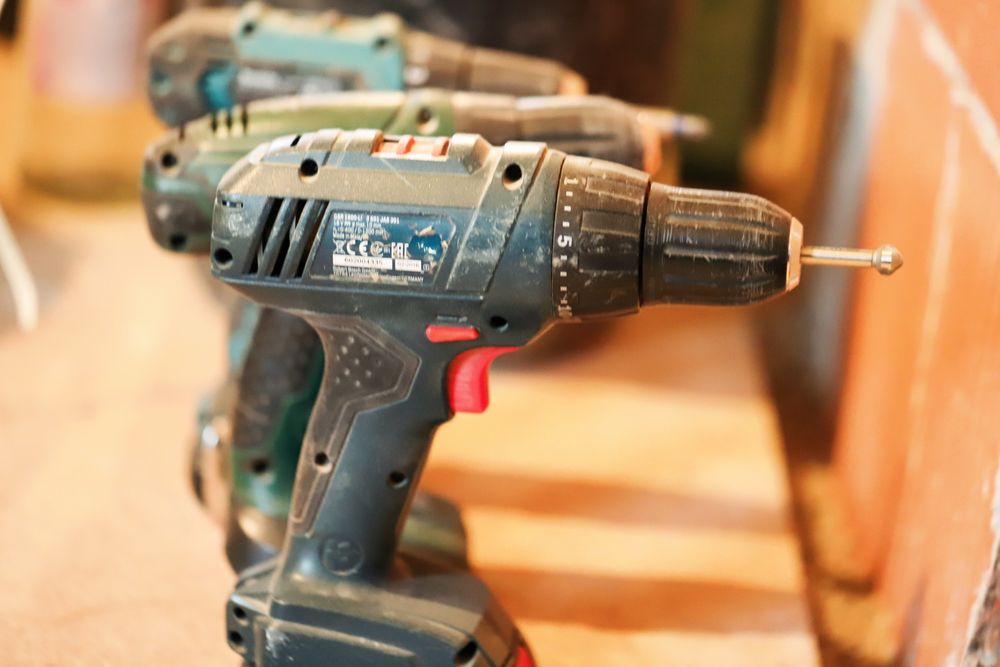 Close-up of a well-used cordless drill with a nail in its chuck, with more drills blurred behind it, against warm wood.