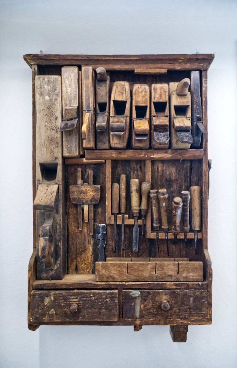 A weathered wooden wall cabinet holding old wooden hand planes, chisels and a marking gauge, with two small drawers underneath, against a plain pale wall.