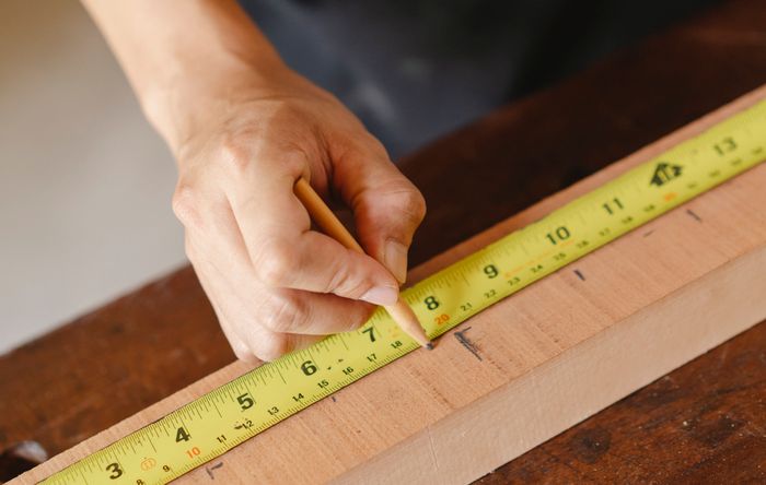 Close-up of a hand marking a lumber plank with a pencil against a yellow tape measure.