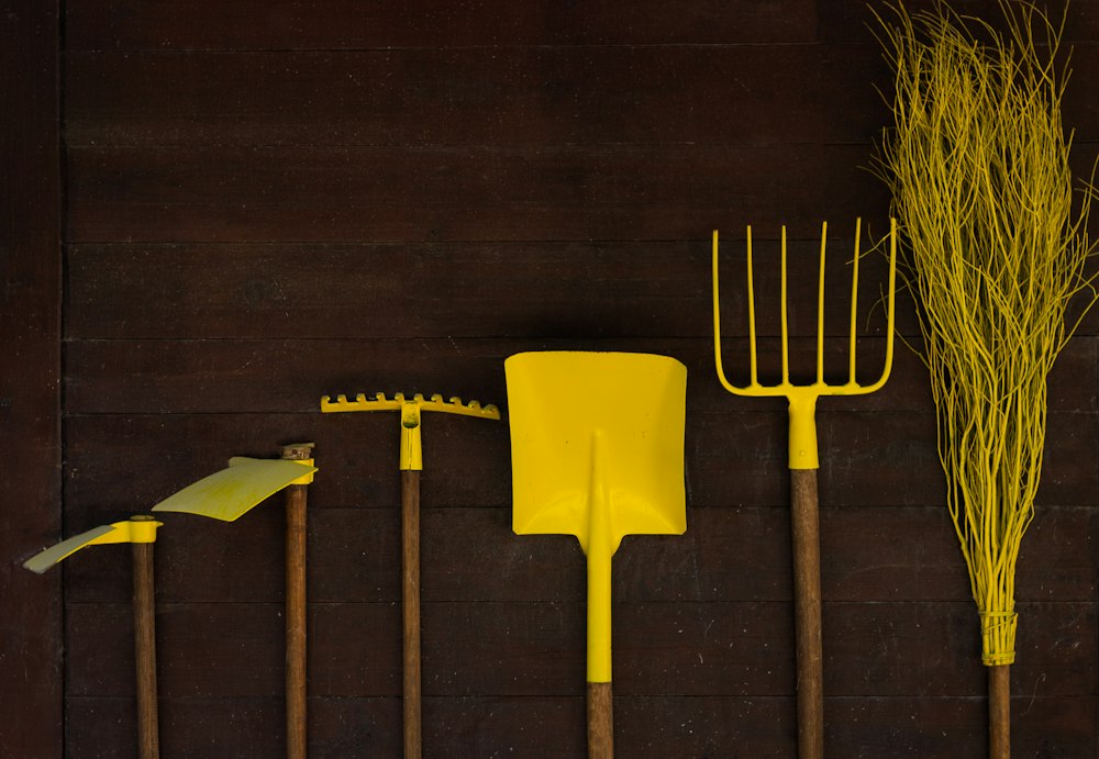 A row of yellow-painted garden tools (hoes, a rake, a shovel, a pitchfork and a twig broom) standing against a dark wooden wall.
