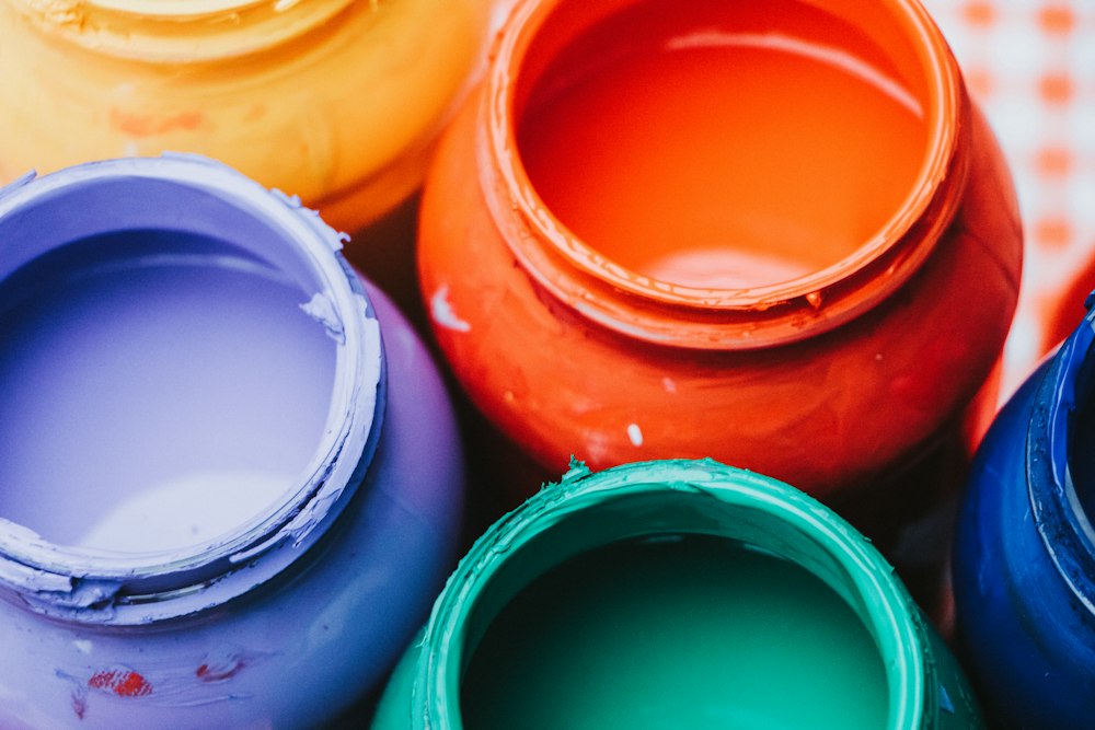 Close-up from above of open tins of paint in lavender, orange-red, teal, yellow and deep blue.