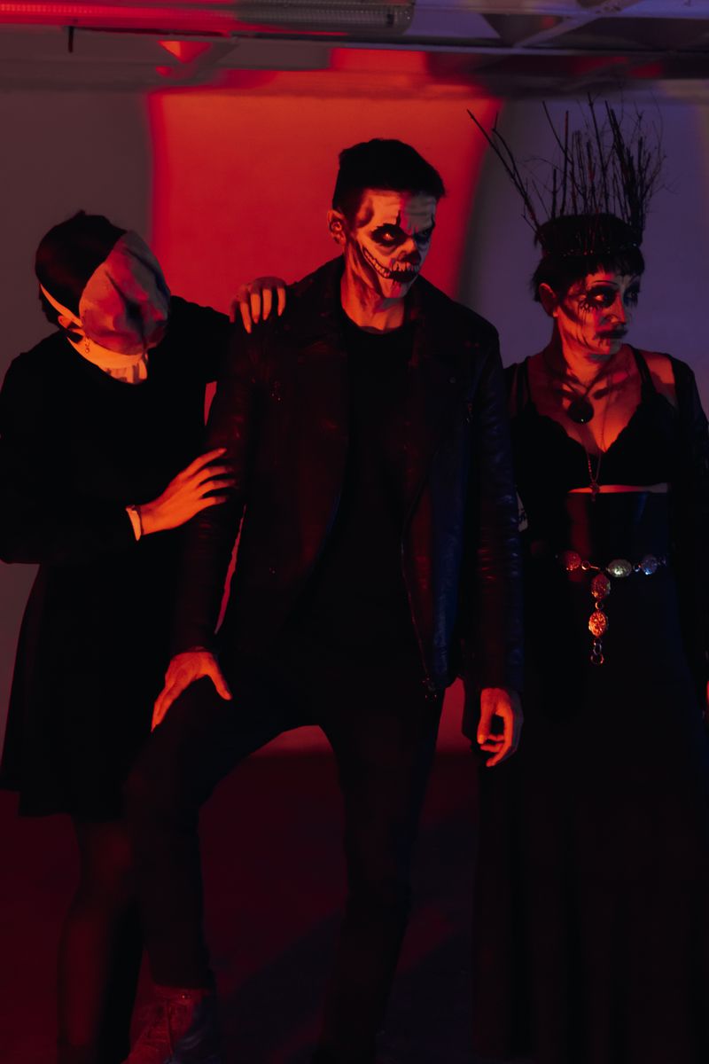 Three adults in dark Halloween costumes stand close together in a room washed in red light: one with a pale cloth mask, a man in skull-style face paint and a leather jacket, and a woman in a twig crown and dark eye makeup.