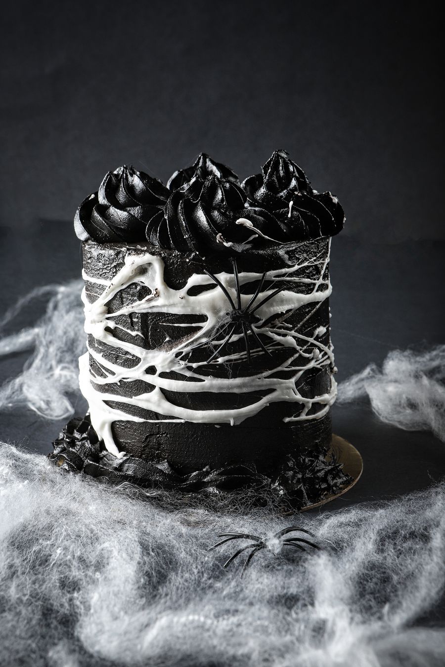 A tall black buttercream cake with black rosettes on top, drizzled with white spiderweb icing and a plastic spider, standing in drifts of fake cobweb on a dark slate surface.