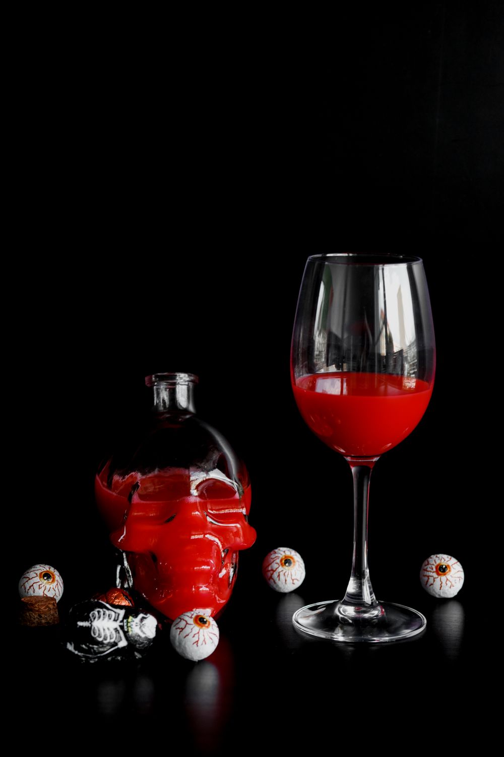 A wine glass of bright red drink beside a glossy red skull-shaped bottle and scattered candy eyeballs on a black background.